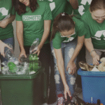 Diverse group of volunteers wearing matching green t-shirts with recycling symbols and slogans like 'ECOMOTTO', smiling and actively sorting plastic bottles, glass, and paper into green and blue bins during a community recycling drive, emphasizing teamwork and environmental awareness.